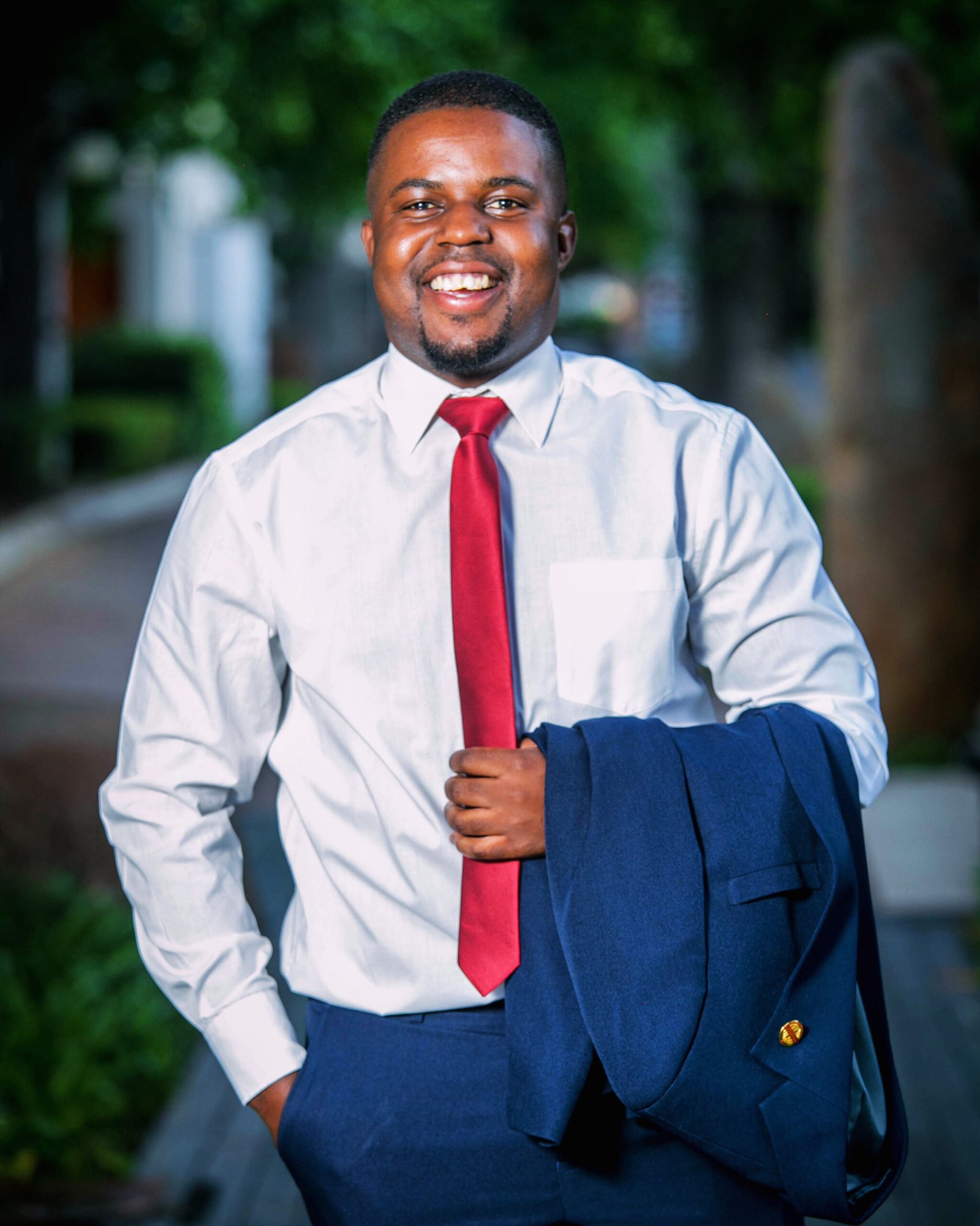 Andrew Tsuro in a white shirt and red tie, holding a navy jacket outdoors.